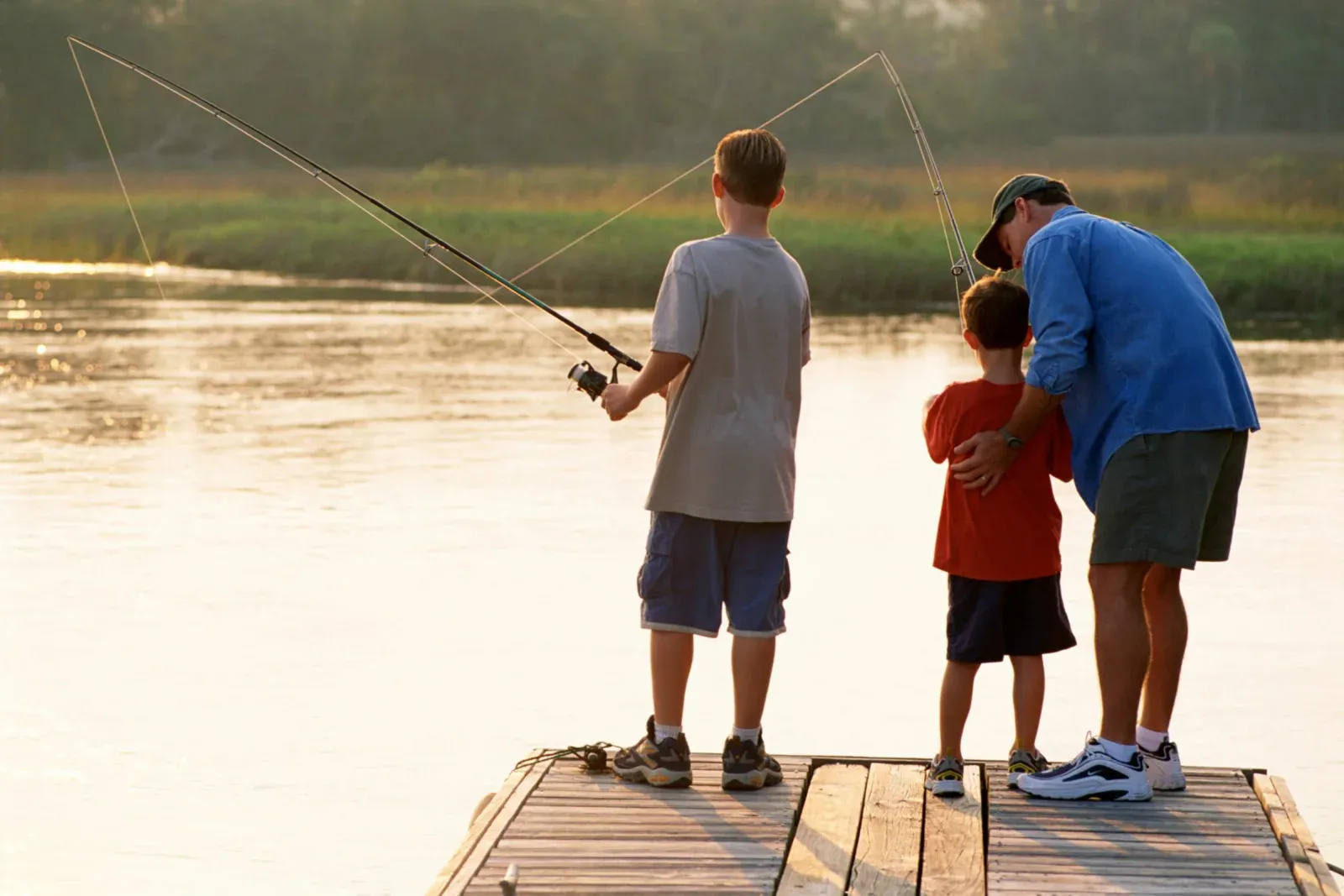 A father figure helping two children fish from a lakeside jetty